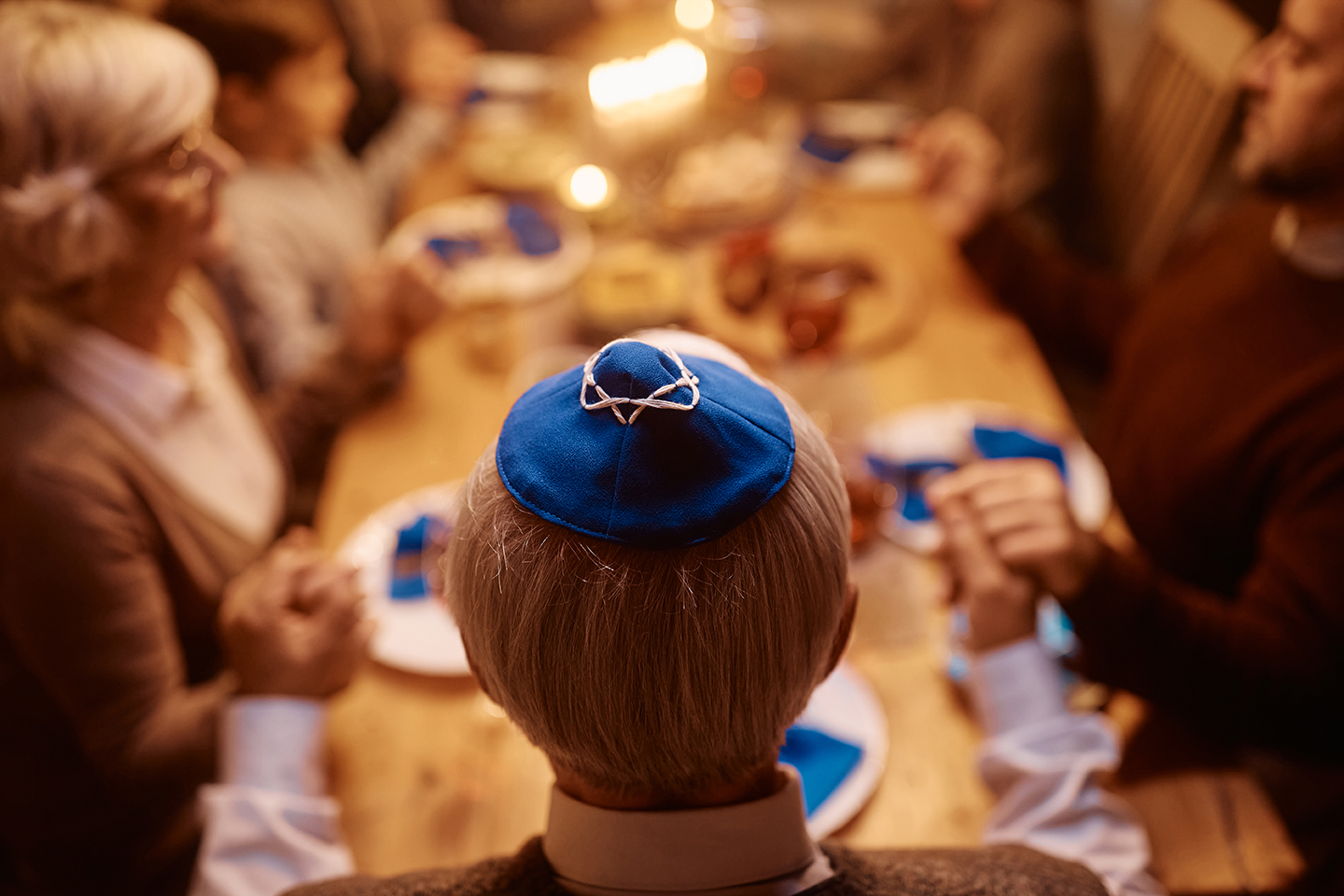 Back view of mature Jewish man and his extended family holding hands and praying before the meal at dining table on Hanukkah.