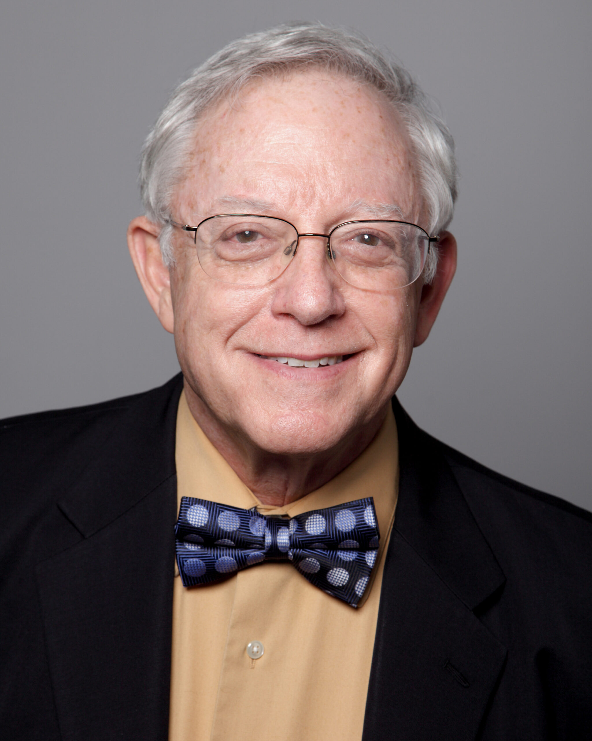 A smiling older man with short gray hair and glasses poses against a plain gray background. He is wearing a tan dress shirt, a patterned navy bow tie with circular designs, and a dark blazer.