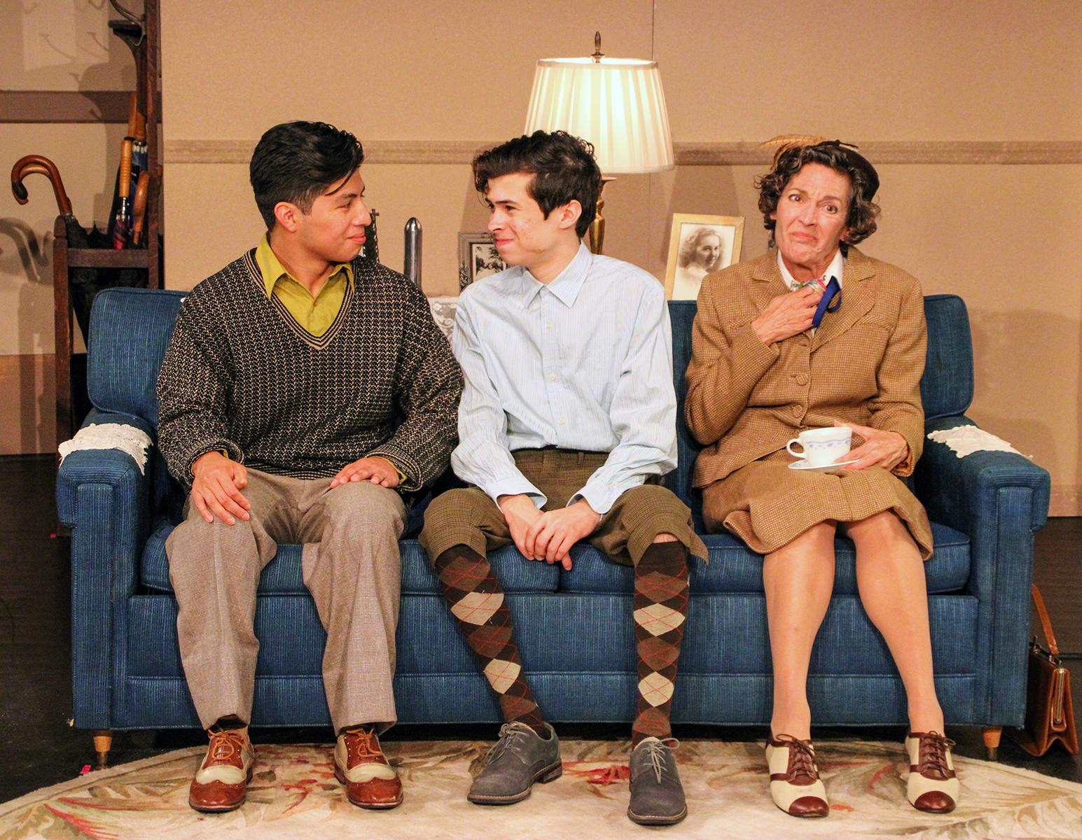 Alt text: Three actors sit closely together on a blue couch in a vintage living room set. The person on the left wears a dark patterned sweater over a green collared shirt, the one in the middle wears a light blue button-down with argyle socks and knicker-style pants, and the woman on the right wears a brown suit, holding a teacup and saucer with a wistful expression. A lamp and framed photograph sit behind them, creating a mid-20th-century domestic scene.