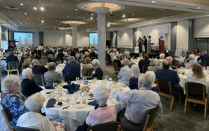 Guests seated at round tables during a Sarasota Jewish Theatre luncheon event, with presenters speaking on stage at the front of the room.