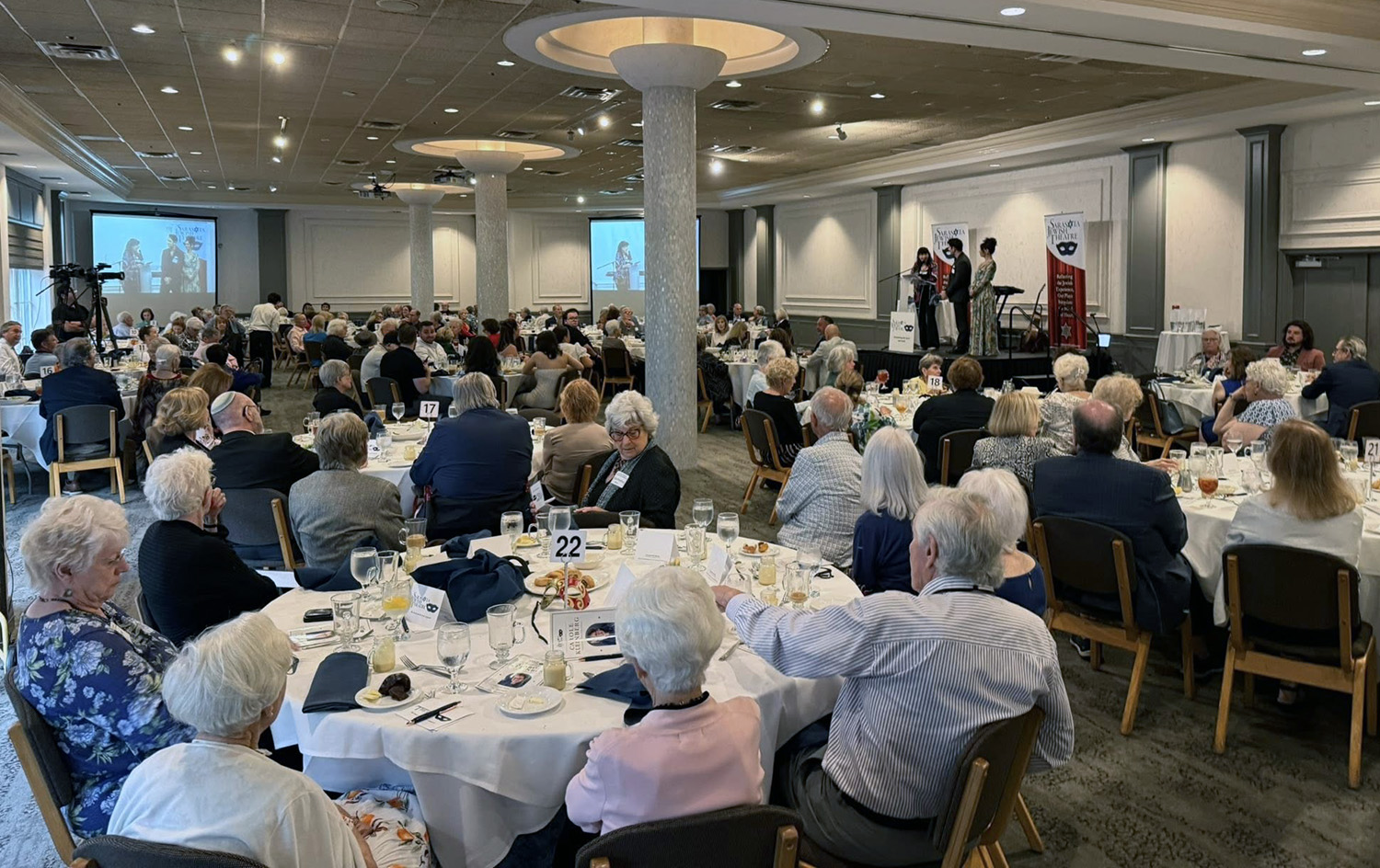 Guests seated at round tables during a Sarasota Jewish Theatre luncheon event, with presenters speaking on stage at the front of the room.