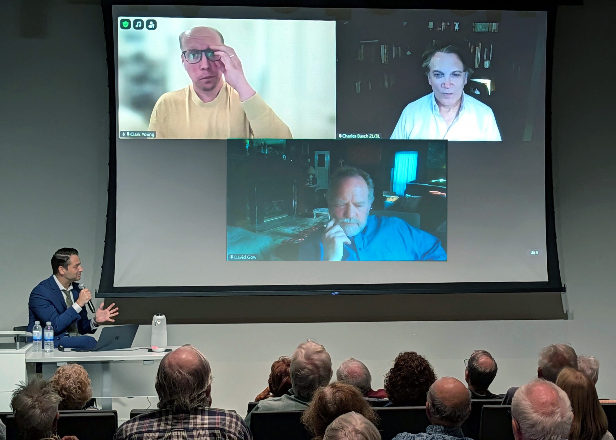 A speaker addresses an audience during a community conversation event, with three virtual panelists displayed on a large screen behind him.