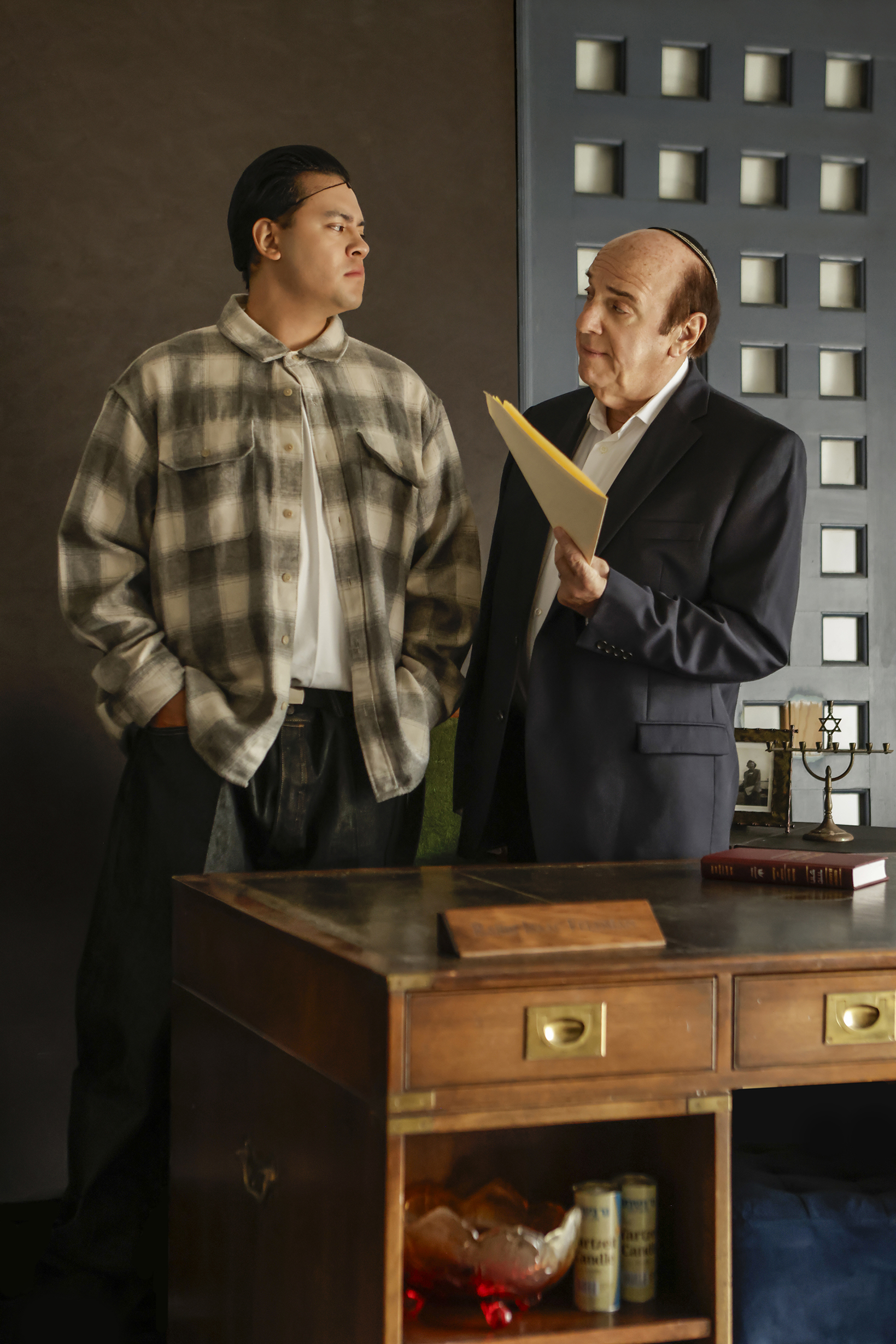 09-29-26_SorchaAugustine_092343 -FINAL Two actors stand beside a desk in conversation, one holding a folder, in Kaddish for the Barrio.