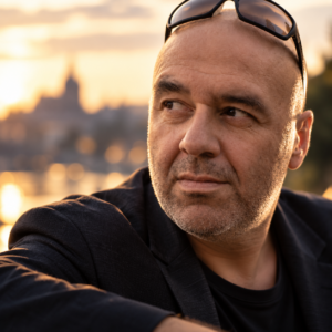 Portrait of Igor Turin wearing sunglasses on his head and a dark jacket, looking to the side against a softly blurred waterfront skyline at sunset.