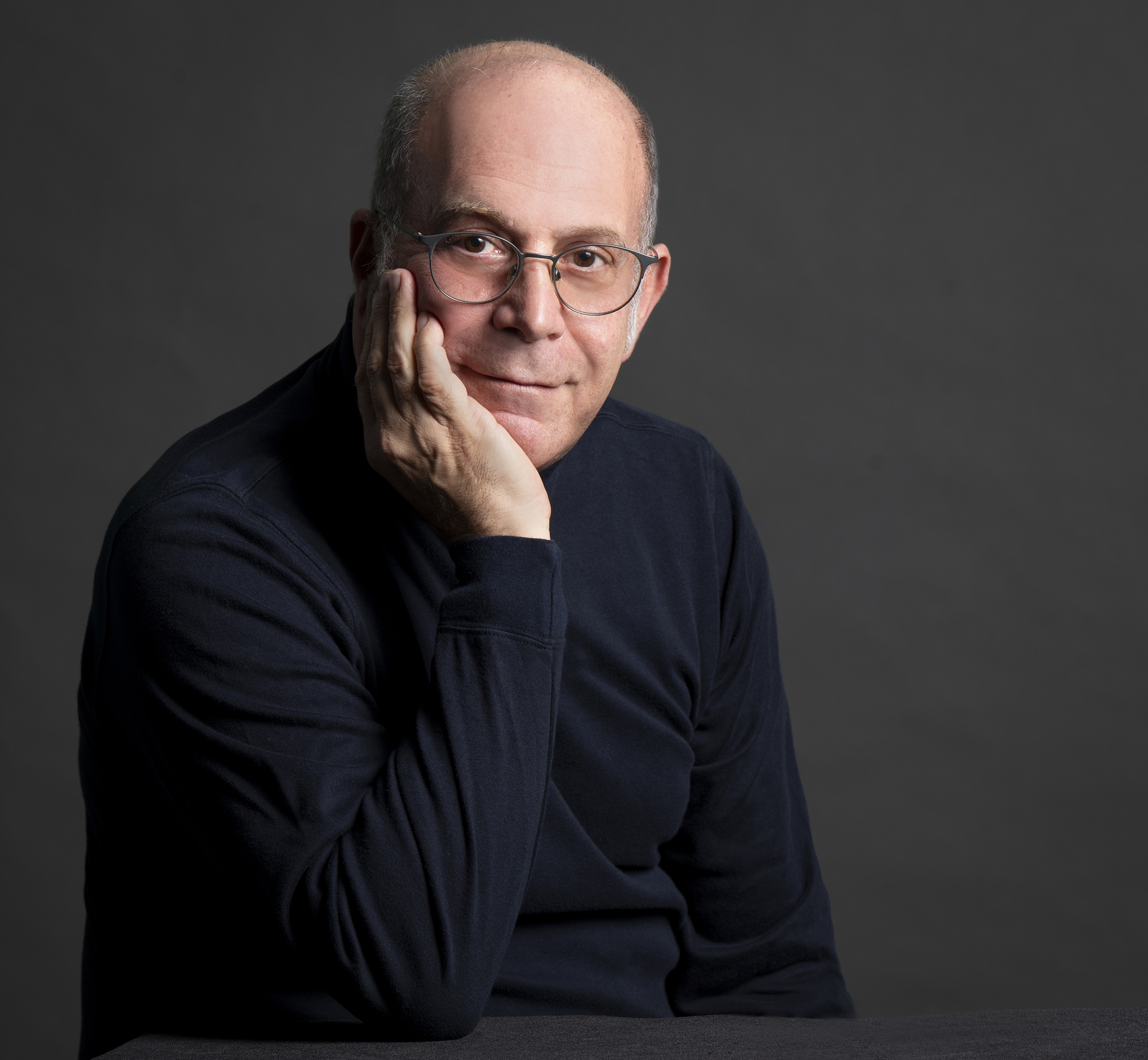 Portrait of Jay Handelman wearing glasses and a dark sweater, resting his face on one hand against a gray background.